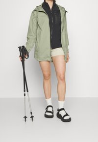 Persoon gekleed in een groene jas, zwarte vest met rits, beige shorts, witte sokken en zwarte sandalen, met in beide handen twee zwarte wandelstokken met metalen punten.