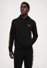 Zwarte hoodie gemaakt van zacht materiaal, met een voorzak en een gouden EA7-logo op de borst en mouw. Klassieke pasvorm met geribbelde manchetten.