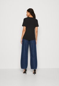 Zwarte tshirt met korte mouwen gecombineerd met blauwe spijkerbroeken met wijde pijpen en zwarte haksandalen, van achteren weergegeven tegen een lichte achtergrond.