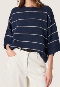 Marineblauer Strickpullover mit weißen horizontalen Streifen, weiten kurzen Ärmeln und einem gerippten Halsausschnitt, kombiniert mit hellen Hosen und einem schwarzen Gürtel.