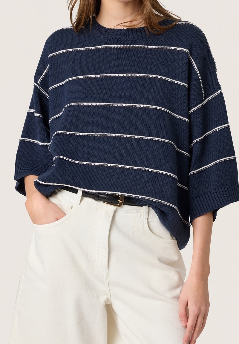 Marineblauer Strickpullover mit weißen horizontalen Streifen, weiten kurzen Ärmeln und einem gerippten Halsausschnitt, kombiniert mit hellen Hosen und einem schwarzen Gürtel.