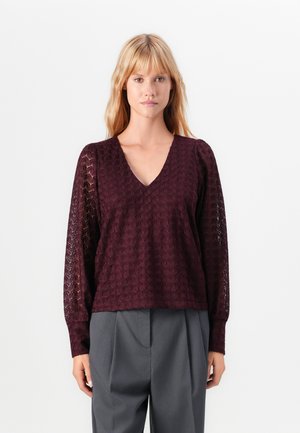 Burgundy textured sweater with long, sheer patterned sleeves and a V-neckline, paired with gray tailored trousers.