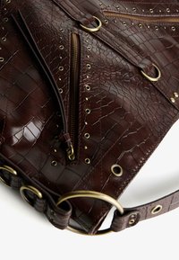Smeđa torba od kože s uzorkom krokodila koja sadrži zlatne hardverske detalje, višestruke zip džepove i kružne akcentne detalje na remenu.