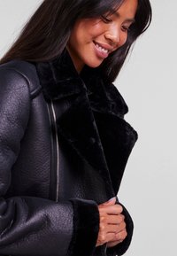 Black faux leather jacket with a plush collar and cuffs, featuring a zip closure and textured finish. Soft, warm texture and structured design.