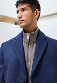Blazer de lana navy superpuesto a un pulóver de cuello alto color topo con un cierre plateado. El blazer cuenta con un cuello estructurado y cierre de botón.