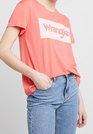 Koraalrood katoenen T-shirt met een wit rechthoekig "Wrangler"-logo. Gecombineerd met lichtblauwe high-waisted jeans, wat een casual pasvorm laat zien.