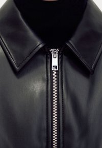 Veste en cuir noire avec un col pointu, dotée d'une fermeture éclair argentée et d'une surface texturée. Vue en gros plan mettant en valeur le détail de la fermeture éclair.