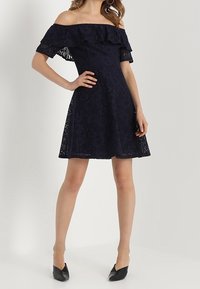 Žena v námořnicky modrých krajkových šatech s odhalenými rameny a volánkovým detailem, ve kterých stojí se jednou rukou na bocích a v černých lodičkách s špičatou špičkou.