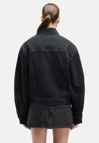 Osoba z brązowym kucykiem, nosząca oversize'ową czarną kurtkę jeansową i pasującą czarną mini spódniczkę jeansową, pokazana od tyłu na białym tle.