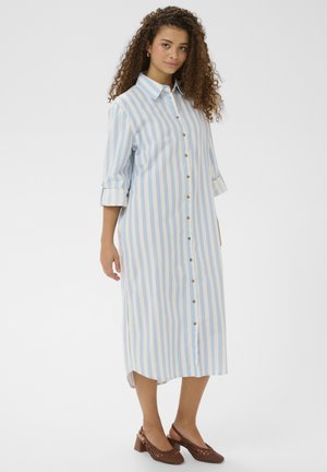 Kvinde iført en midi-skjortekjole med blå og hvide lodrette striber og brune lave hælede flettede sko, stående foran en ensfarvet baggrund.