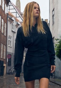Černé pletené šaty s límcem na knoflíky, dlouhými rukávy a přiléhavým střihem. Materiál vypadá měkký s texturovaným povrchem.