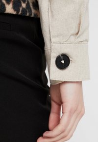 Beżowa lniana mankiet z czarnym detalem guzika. Mankiet ma czyste wykończenie z subtelną teksturą, w połączeniu z czarną odzieżą.