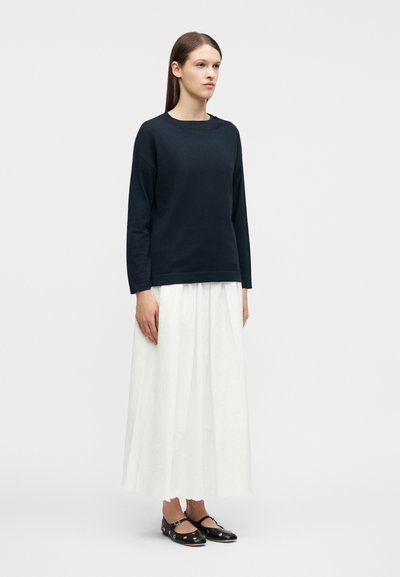 Γυναίκα που στέκεται φορώντας μακρύ λευκό φούστα, μαύρο μακρυμάνικο τοπ και μαύρα παπούτσια Mary Jane με ασημένιες τρύπες σε απλό φόντο.