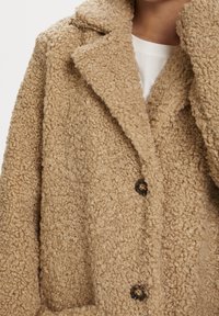 Abrigo de piel sintética marrón claro, texturizado, con cuello con solapa, cierre de botones y dos bolsillos laterales. Material suave y esponjoso.