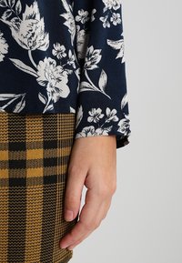 Námořnická blůza s bílým květinovým vzorem, zkombinovaná s kostkovanou sukní v černé a zlaté barvě. Detailní záběr na ruku odpočívající na okraji sukně.