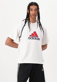 Hvid bomulds t-shirt med korte ærmer, har et rødt og marineblåt Adidas-logo på brystet, kontrasterende syninger langs skuldersømmen.