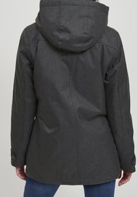 Person som bär en mörkgrå huva-jacka sedd bakifrån, i kombination med blåjeans, stående mot en enfärgad bakgrund.