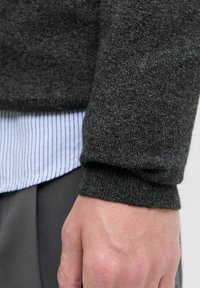 Gros plan sur le poignet d'une personne portant un pull en tricot gris foncé par-dessus une chemise à rayures bleu clair, avec un pantalon gris visible en dessous.
