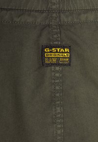 G-Star Originals -merkki tumman vihreällä kankaalla, jossa on keltaista ja mustaa tekstiä. Ompeleet näkyvät sileällä pinnalla, jossa on näkyviä sauman yksityiskohtia.