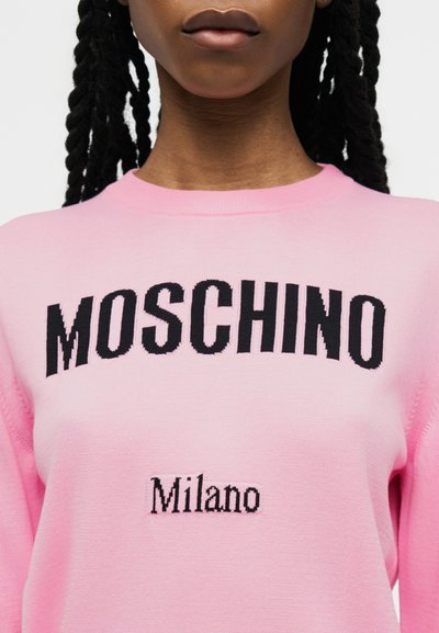 Gros plan sur une personne portant un pull rose avec le texte noir "MOSCHINO" et plus petit "Milano" en dessous à l'avant.
