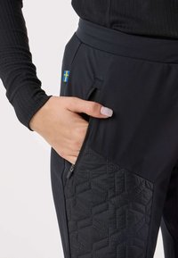 Pantaloni atletici neri con tasca con cerniera e motivo geometrico trapuntato sul lato. Il materiale è liscio con una leggera lucentezza.