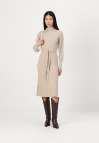 Robe en tricot côtelé beige avec un col haut et une taille ceinturée, associée à des bottes de cowboy marron foncé jusqu'aux genoux. Texture lisse et longueur mi-mollet.