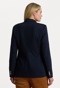 Donna con capelli biondi che indossa un blaser blu navy con bottoni dorati e pantaloni in velluto a coste marroni, mostrata di spalle su uno sfondo bianco.