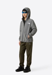 Sivá mikina na zips s kapucňou, pod ňou čierne tričko, zelené športové nohavice a čierne topánky. Slnečné okuliare s modrým tónovaným sklom.