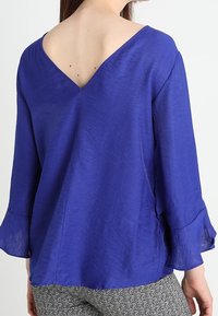 Blusa leggera viola con scollo a V e maniche a campana, caratterizzata da una sottile texture e un design con schiena aperta.