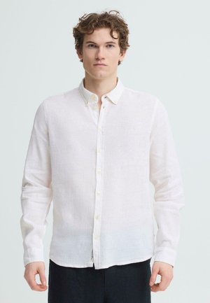 Młody mężczyzna o kręconych włosach, ubrany w białą, zapinaną na guziki koszulę z długim rękawem i ciemne spodnie, twarzą zwrócony do przodu na jasnym, jednolitym tle.