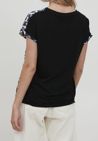 Camiseta negra de manga corta con un acento de estampado de leopardo en los hombros. Tejido suave, escote redondeado y corte holgado. Combinada con pantalones blancos.