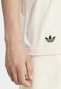 Vit bomulls-t-shirt med svart broderad logotyp nära fållen. Mjuk textur, avslappnad passform, kombinerad med ljusfärgade shorts. Inga mönster.