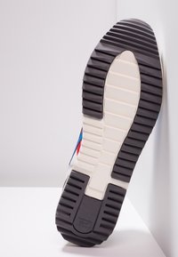 Suola di sneaker con battistrada in gomma nera texturizzata, intersuola beige e dettaglio del logo in evidenza, inquadrata su uno sfondo bianco.