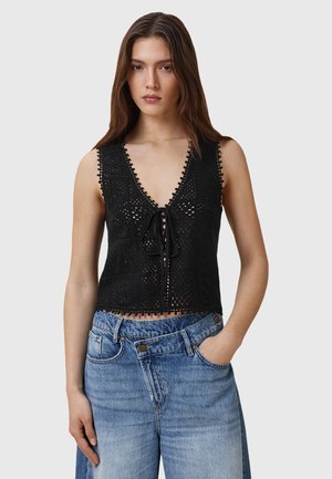 Top corto in crochet nero con scollatura a V profonda, apertura anteriore legata, bordi scallop e motivi intricati. Abbinato a jeans azzurri a vita alta.