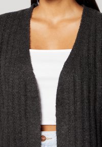 Cardigan lavorato a maglia grigio scuro con texture a coste e apertura frontale, indossato su una canotta bianca aderente, che mostra un look casual a strati.
