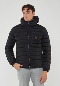 Giacca a vento nera imbottita con motivo a righe orizzontali, colletto alto, zip frontale e una piccola tasca sul petto con patch del logo.