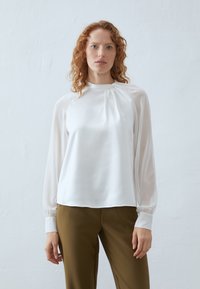 Bílá blůza vyrobená z hedvábné látky, s dlouhými nabíranými rukávy, vysokým výstřihem a sklady na přední straně, doplněná olivově zelenými kalhotami.