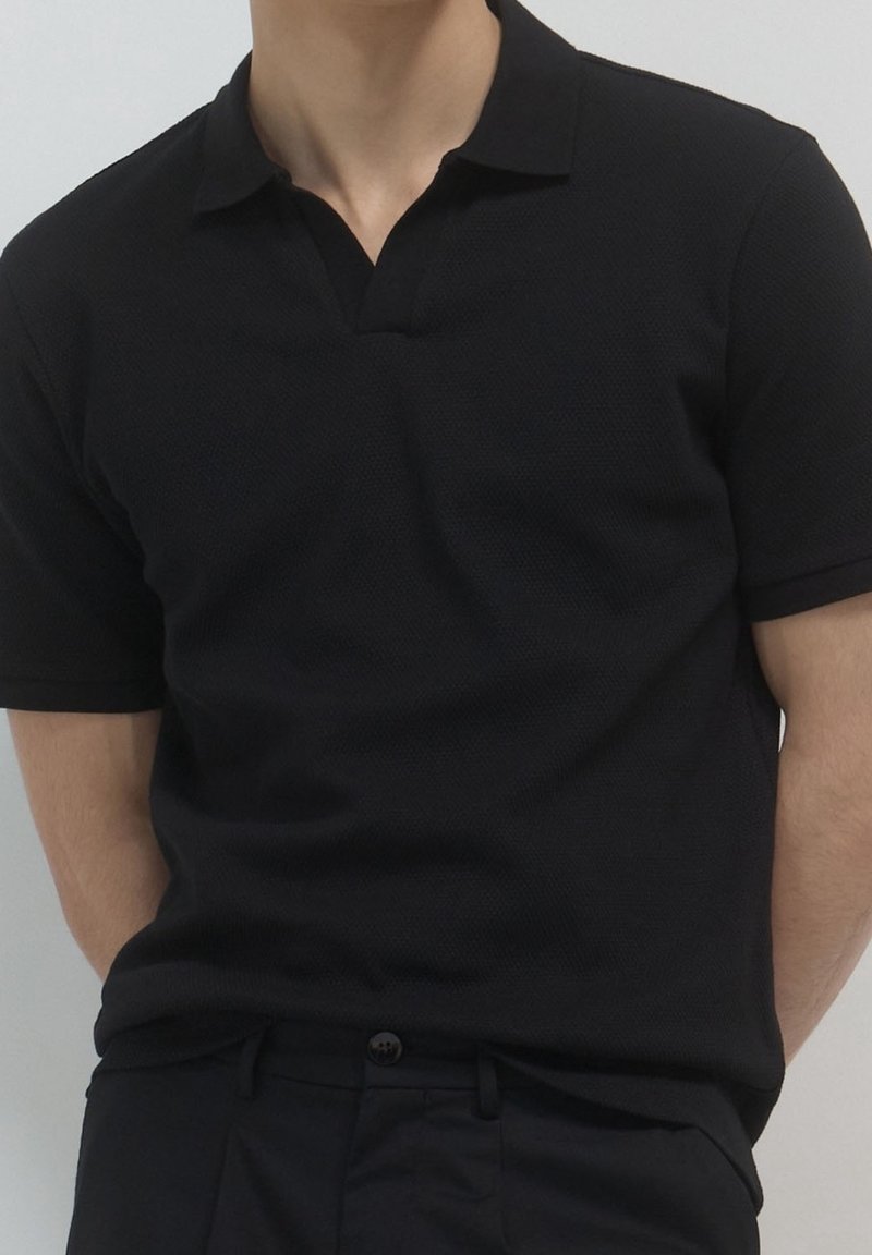 Personne portant un polo noir uni à manches courtes rentré dans un pantalon noir, sur un fond neutre.