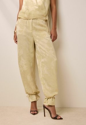 Femeie purtând pantaloni largi bej cu model floral și glezne strânse, asortați cu top potrivit și sandale negre cu toc înalt.