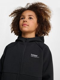 Ung person med krøllet hår iført en sort hættetrøje med lynlås, der kigger let opad mod en ensfarvet baggrund.
