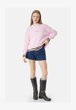Joven mujer con suéter rosa "Angel", camisa de rayas, pantalones cortos vaqueros, calcetines grises y botas negras de cuero hasta la rodilla, de pie con la mano en la cadera.
