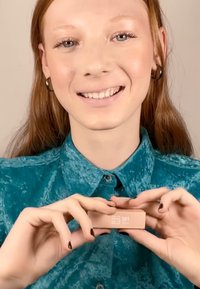 Modellen holder en lys rosa rektangulær boks med en hvit etikett som viser "501." Modellen har på seg en teal, teksturert skjorte med knapper.