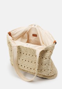 Gebreide beige draagtas van geweven materiaal, met een zachte canvasvoering en een lederen label, voorzien van twee lange handvaten om te dragen.