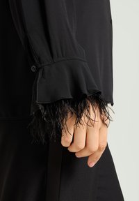 Zwarte blouse met een gerimpelde, gevederde manchetkant. De mouw heeft een gladde stof en knoopdetail bij de pols. Gestructureerde accenten aan de zoom.
