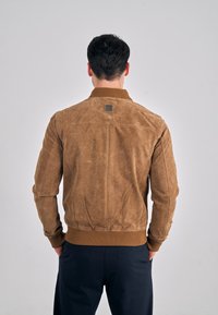 Ruskea mokkainen bomber-takki, jossa on resorikaulus ja -hihansuut, tikattu paneelisuunnittelu sekä logopatches selässä.