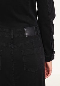 Svart denimjacka som visar ryggdetaljer, inklusive en läderlapp, två bakfickor och kontrasterande sömmar längs sidsömmarna.