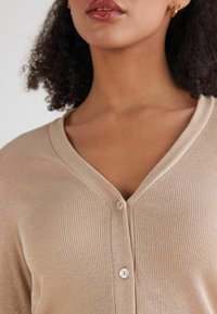 Beżowy kardigan prążkowany z dekoltem w kształcie V, z przodem zapinanym na guziki i subtelnym blaskiem. Miękka tkanina o gładkiej strukturze i lekkim rozciągnięciu.