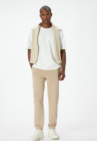 Completo in cotone beige con top senza maniche drappeggiato su una t-shirt bianca, abbinato a pantaloni jogger coordinati e sneakers bianche alta.