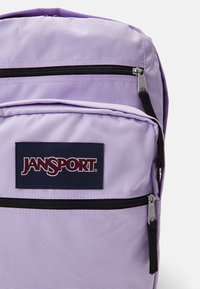 Šviesiai violetinė Jansport kuprinė su dideliu pagrindiniu skyriumi, dviem priekiniais užtrauktukais uždaromais kišenėmis ir ryškiu tamsiai mėlynu logotipo ženklu.