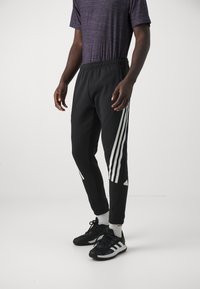 Pantaloni sportivi neri con strisce bianche sui lati e logo. Vita elastica, vestibilità affusolata, realizzati in materiale morbido, indossati con scarpe da ginnastica nere.